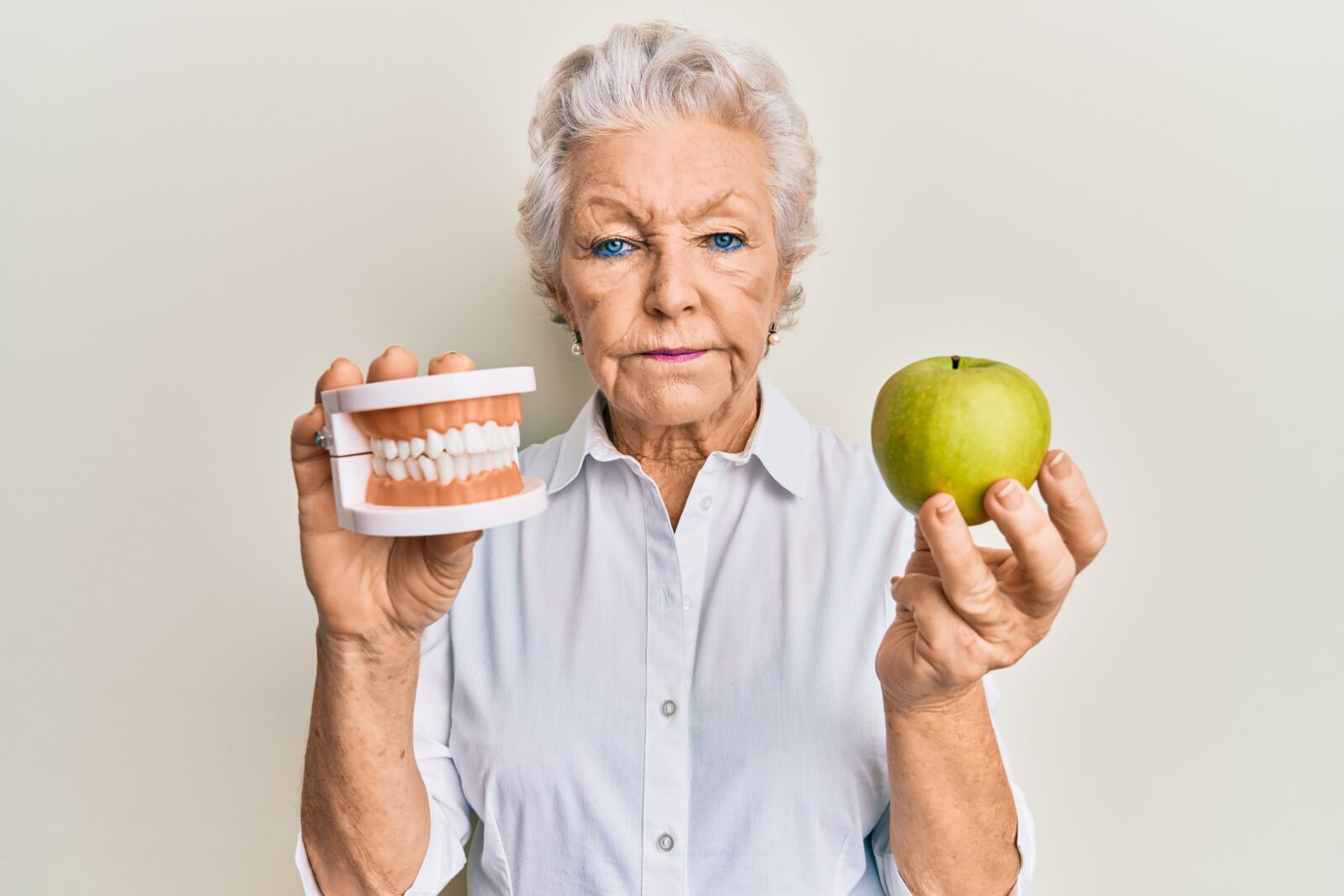 older woman holding dentures in one hand and an apple in the other with an unhappy expression on her face